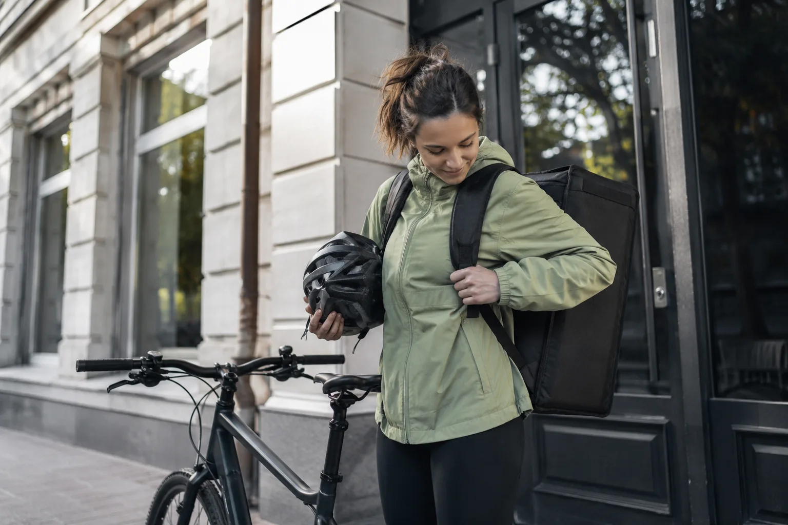Livreuse à vélo ajustant la sangle de son sac de livraison isotherme devant un immeuble, le matin