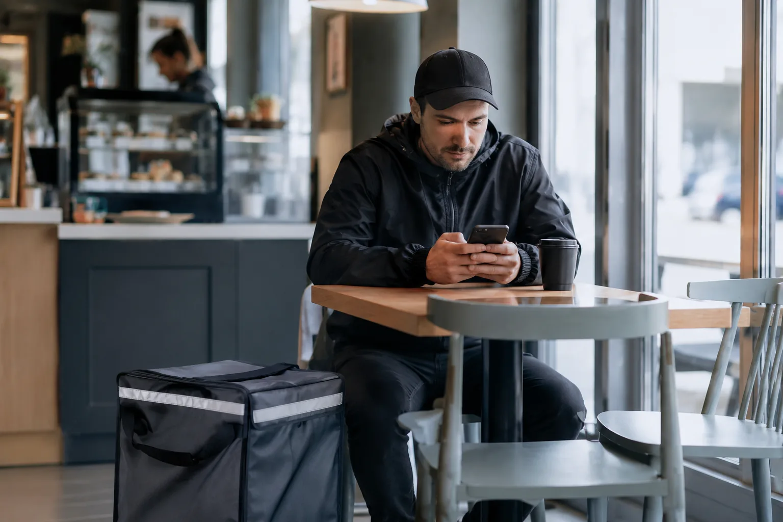 Livreur en pause dans un café, consultant son smartphone, sac de livraison isotherme posé au sol
